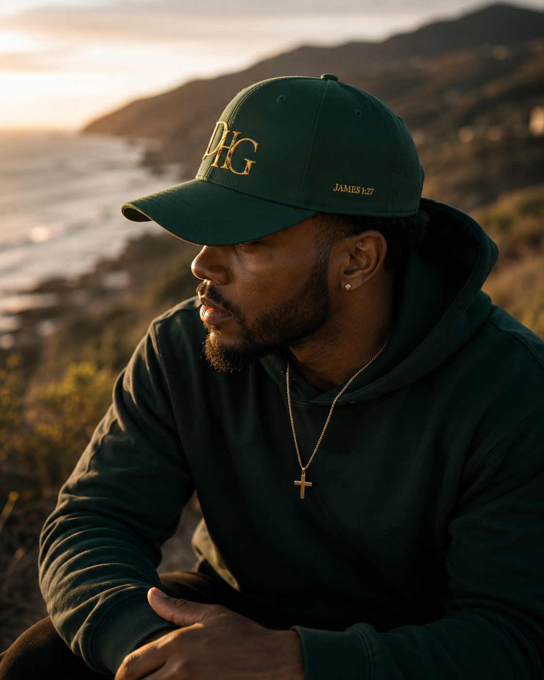 A man wearing a dark green baseball cap with gold embroidered lettering and a matching dark green hoodie, sitting outdoors with a coastal mountain landscape in the background during golden hour.