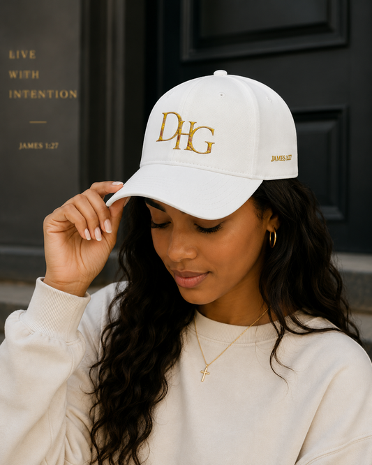A woman wearing a white baseball cap with gold embroidered lettering that reads 'DHG' on the front and 'JAMES 1:27 on the side, paired with a cream sweatshirt.